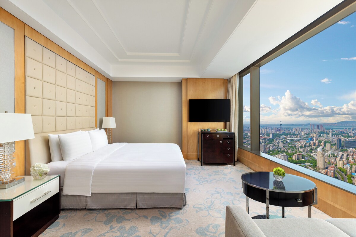Une chambre d’hôtel avec un grand lit, une télévision, une petite table avec une plante et une vue sur la ville à travers de grandes fenêtres.