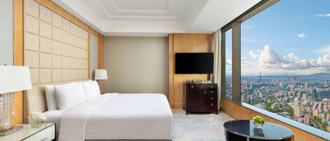 A hotel room with a large bed, a TV, a small table with a plant, and a city view through large windows.