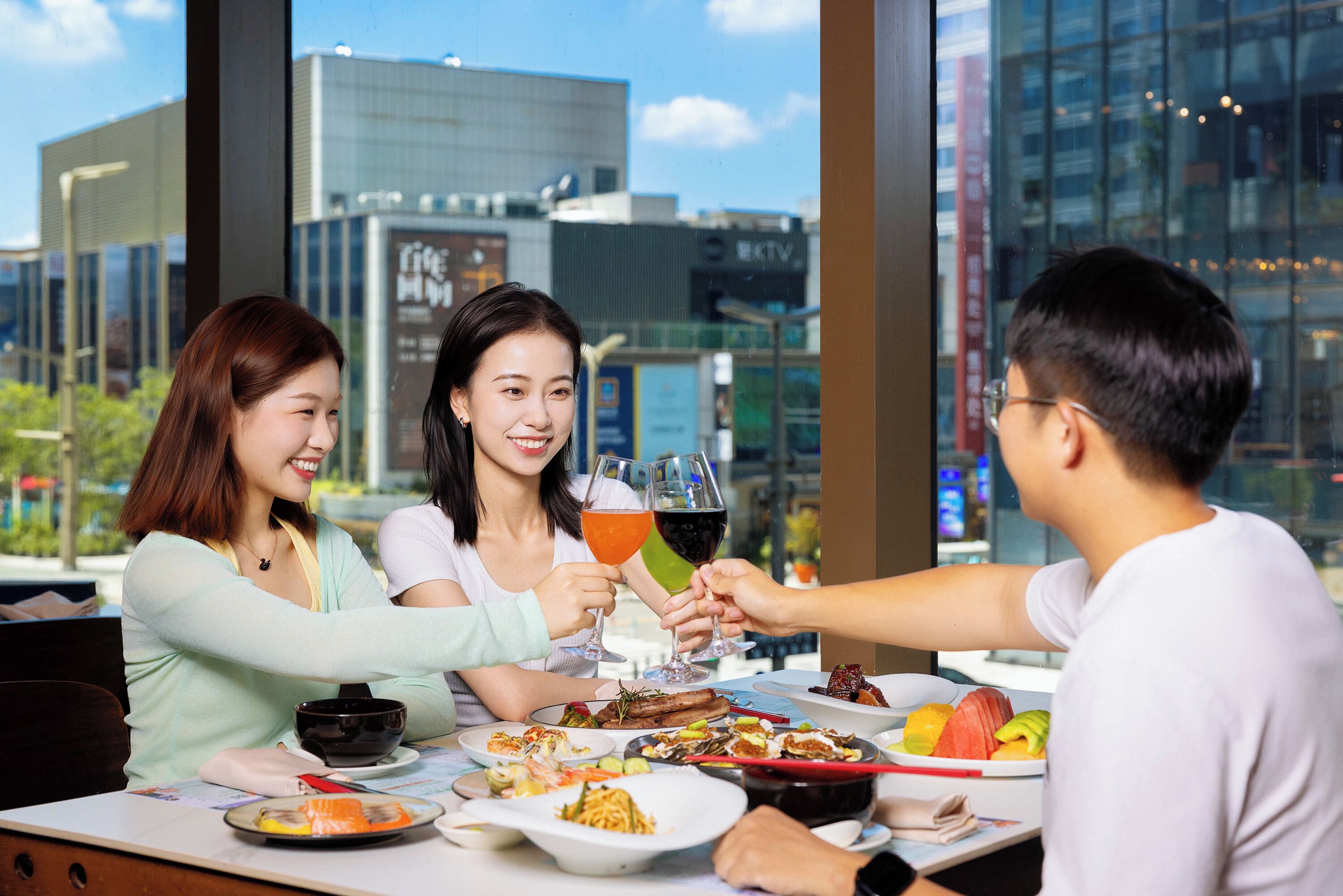 A group of people toasting at a table with various dishes and drinks, with a cityscape view through large windows.