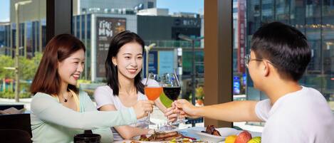 A group of people toasting at a table with various dishes and drinks, with a cityscape view through large windows.