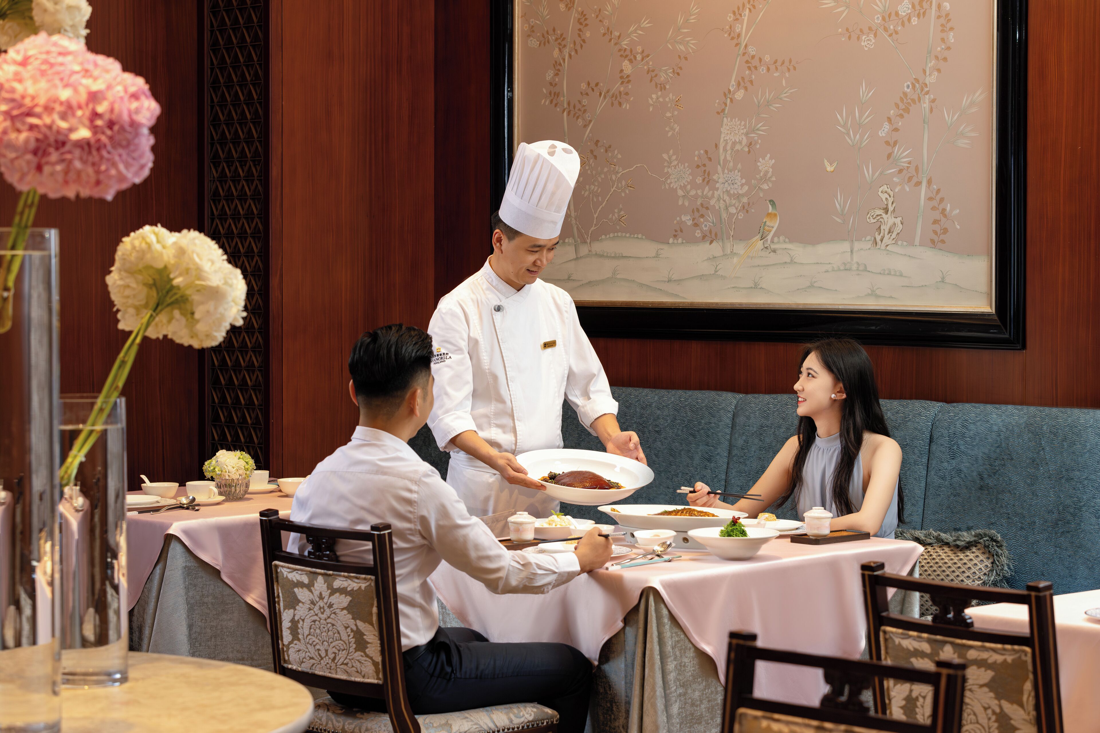 A dining area with a table set for three, a waiter serving food, and a painting on the wall.