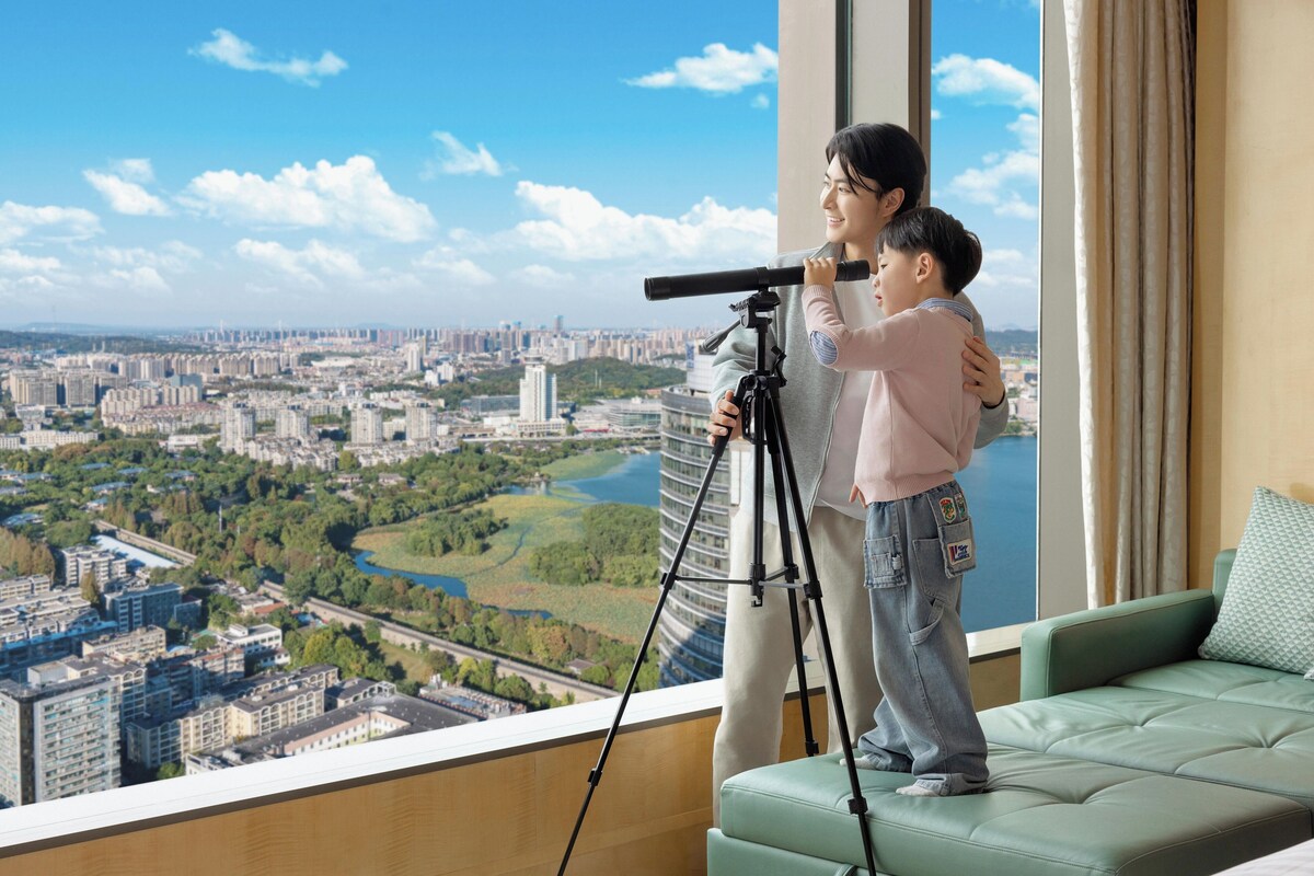 A room with a large window overlooking a cityscape, a telescope on a tripod, and a green sofa.