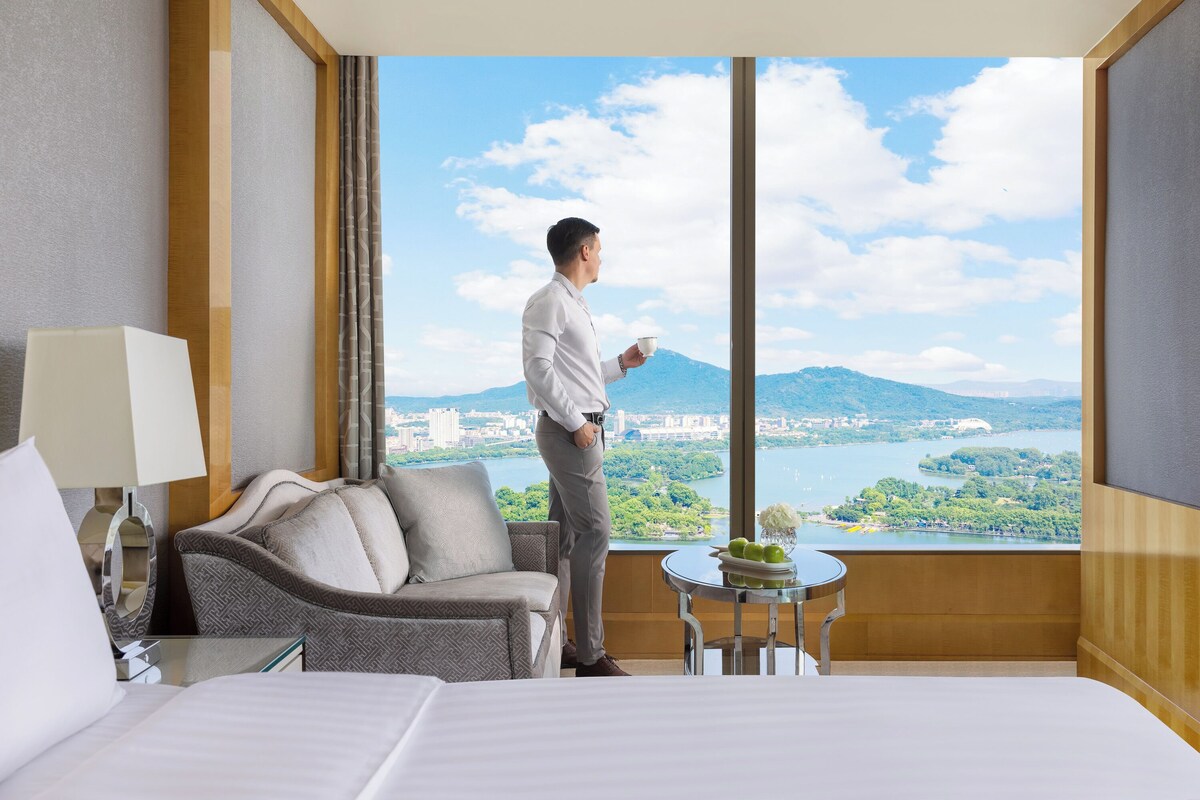 A hotel room with a large window, a bed, a sofa, a small table with fruit, and a lamp.