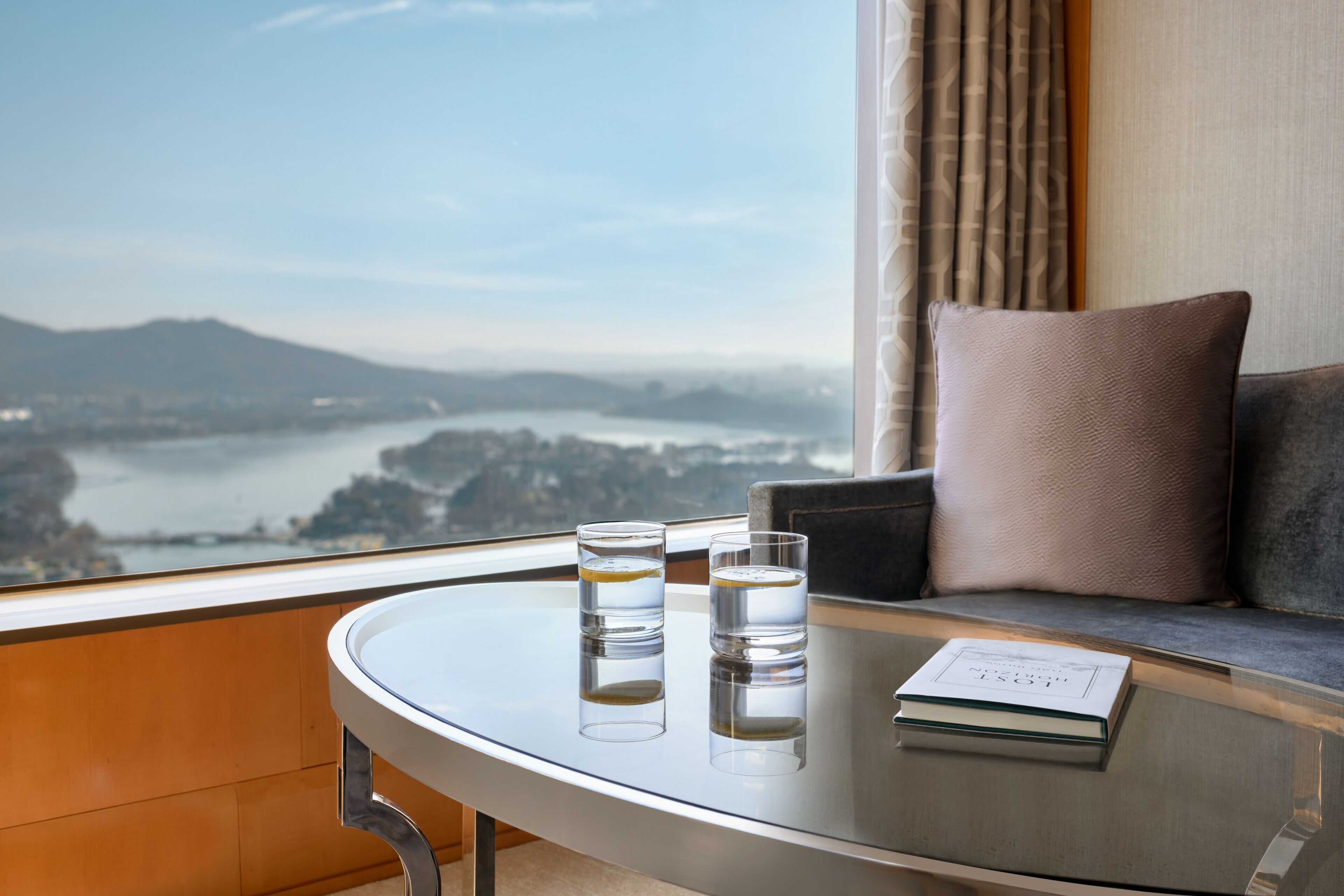 A modern room with a glass table, two glasses of water, a book, and a view of a lake and mountains through a large window.