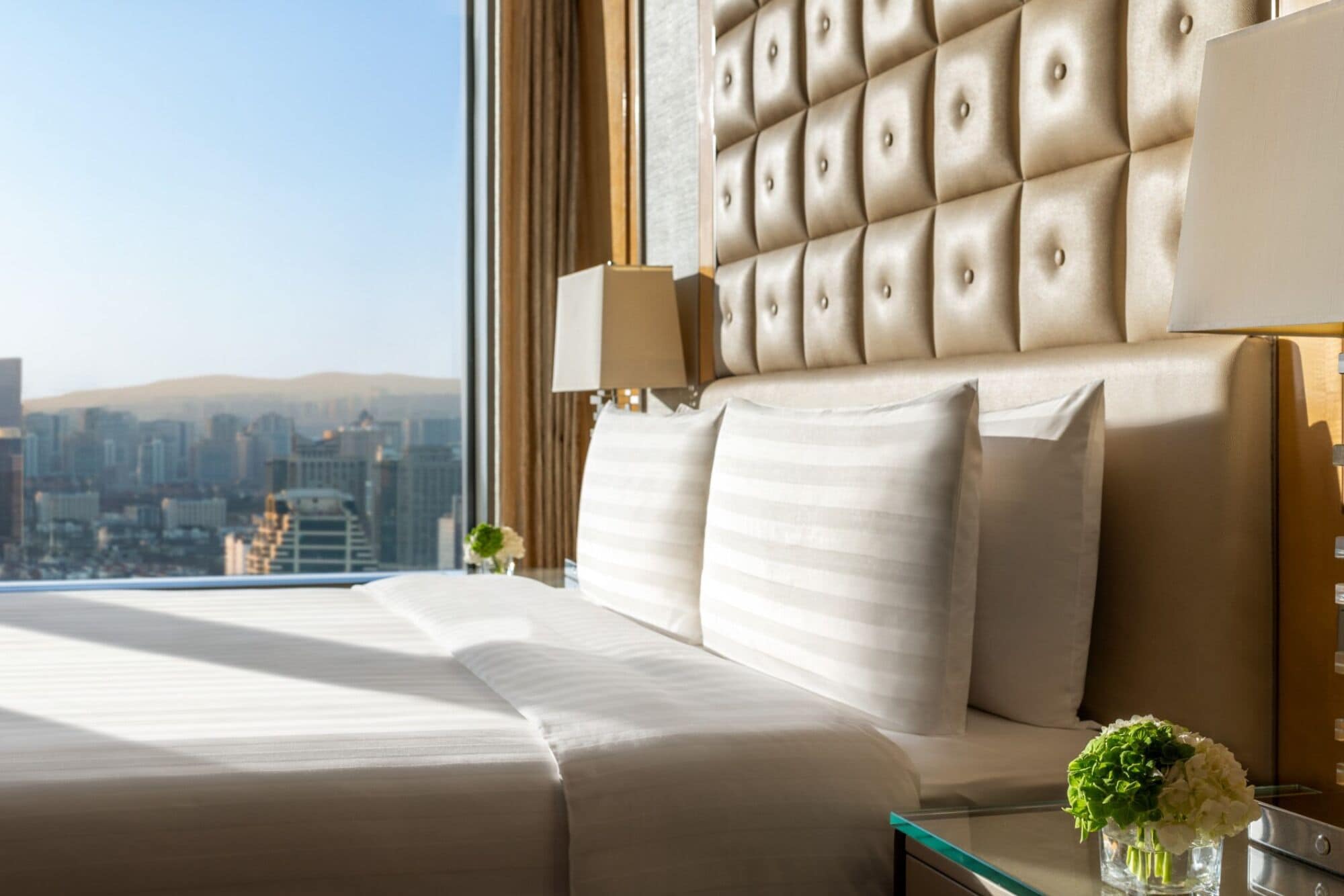 Ein Hotelzimmer mit einem großen Bett, einem gepolsterten Kopfteil, einem Glastisch mit Blumen und Blick auf die Stadt.