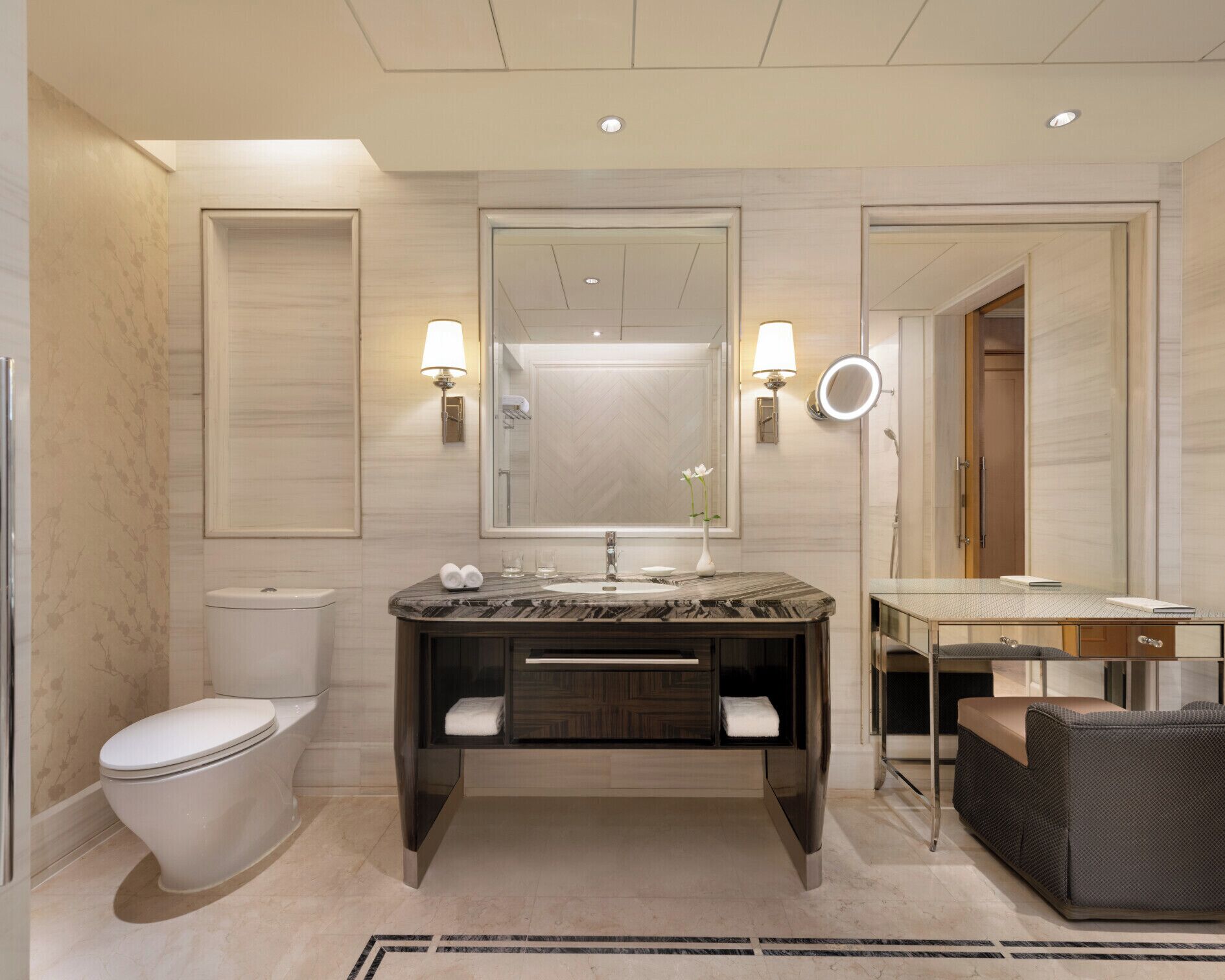 Un baño moderno con lavamanos de mármol, inodoro, espejo con iluminación y una zona de estar con una silla y una mesa.