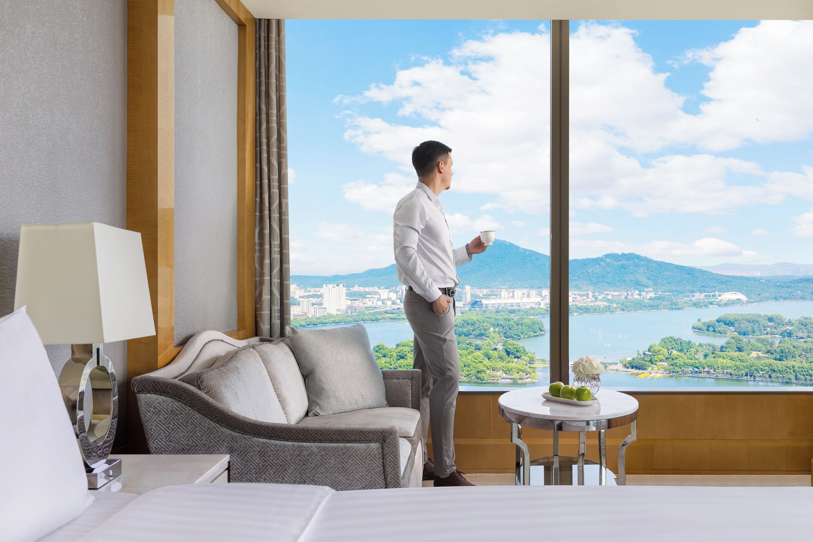 A hotel room with a large window, a sofa, a table with a vase, and a bed.