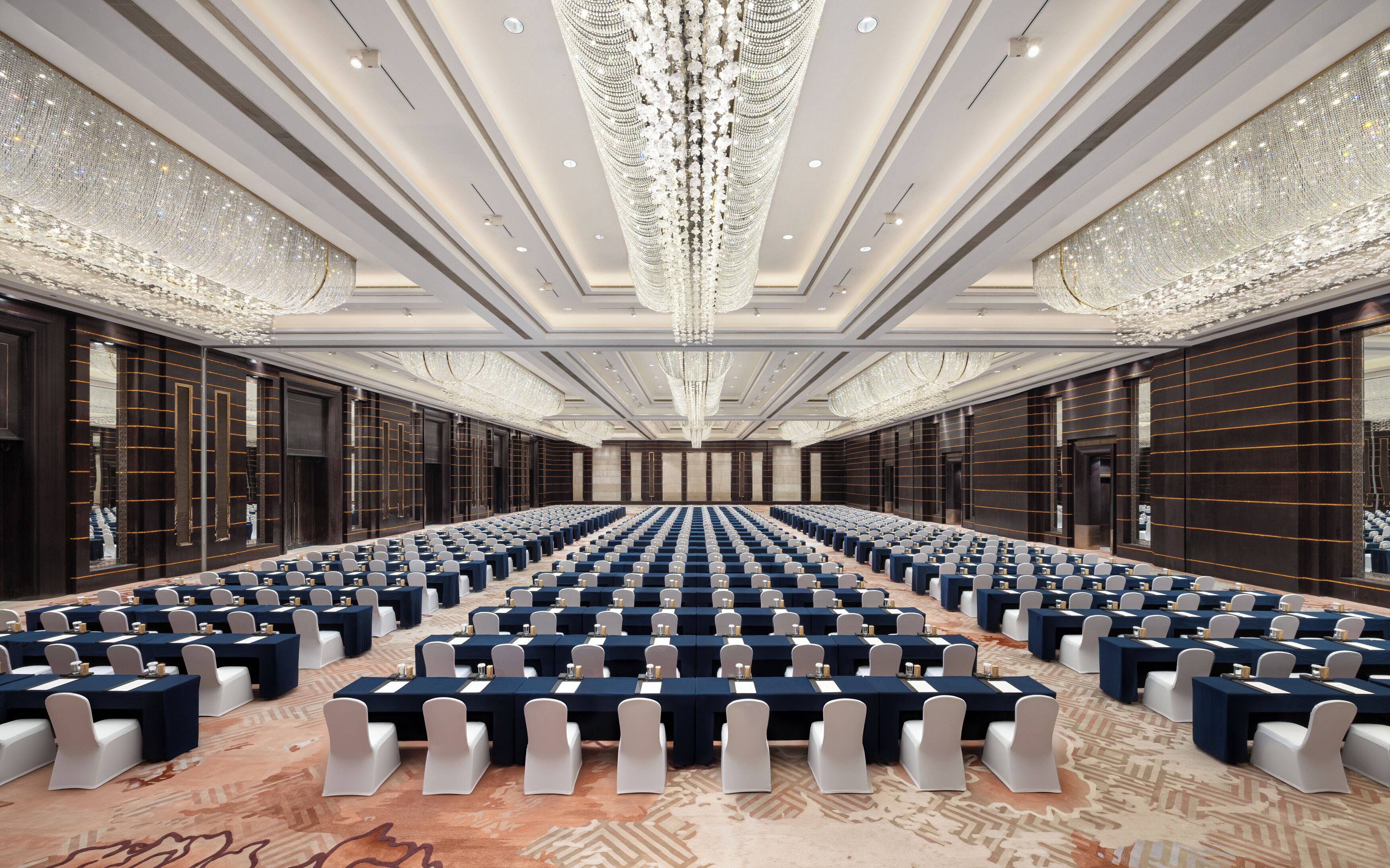 A large conference room with rows of tables and chairs, ornate chandeliers, and a patterned carpet.