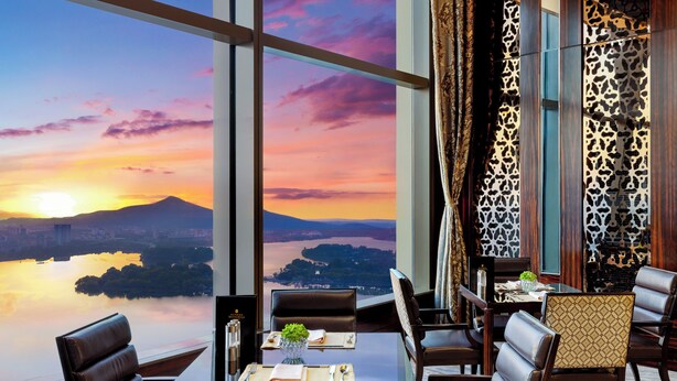 Un comedor con una mesa lista para dos, vista a un atardecer sobre un cuerpo de agua y un panel de madera decorativo en la pared.