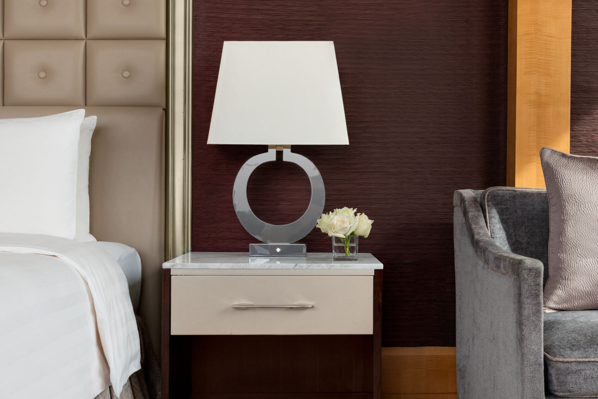 Habitación de hotel con cama, mesita de noche con lámpara, un jarrón con flores y una silla.