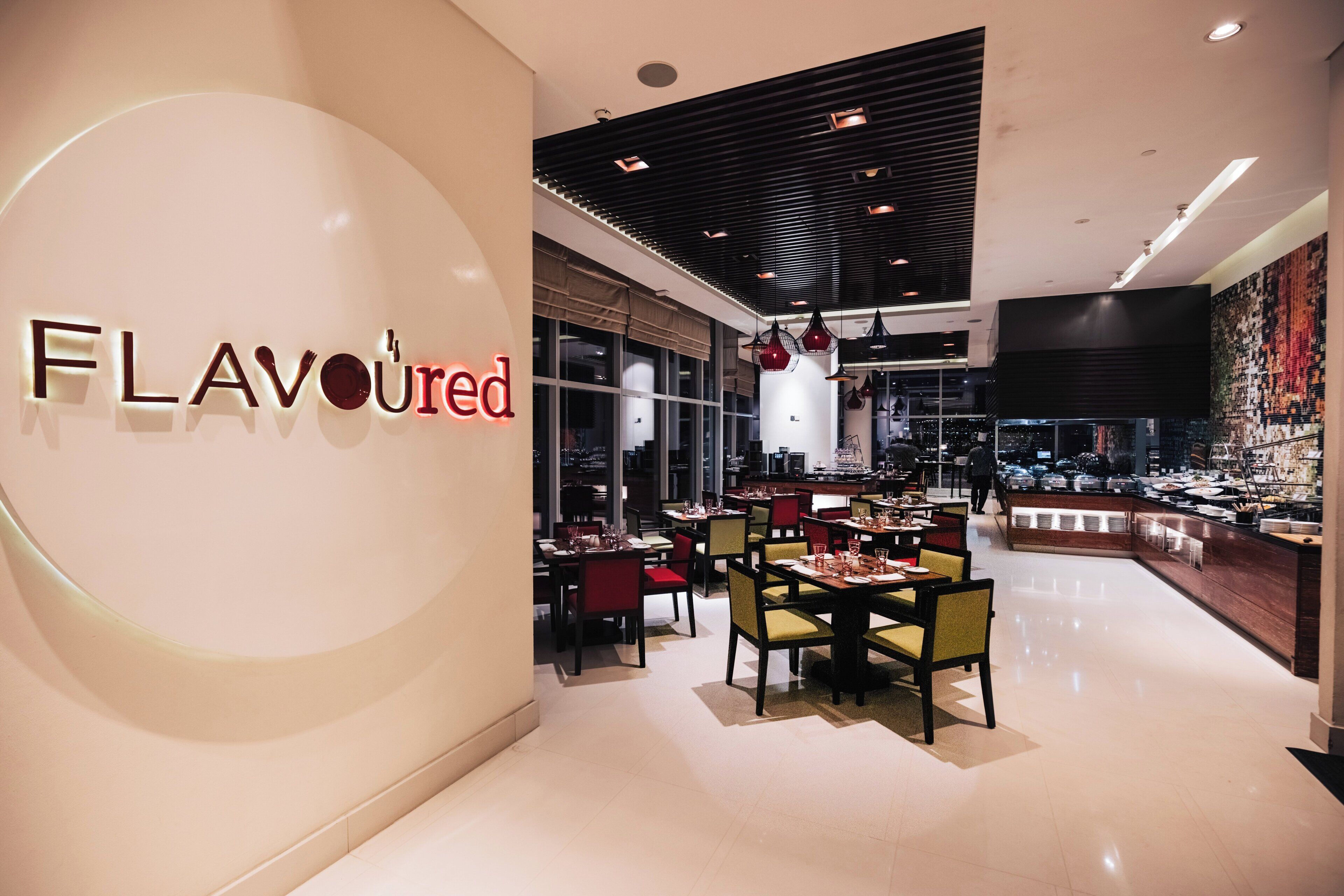 A modern restaurant interior with a prominent 'FLAVOURED' sign, dining tables, chairs, and a kitchen area visible through the glass.