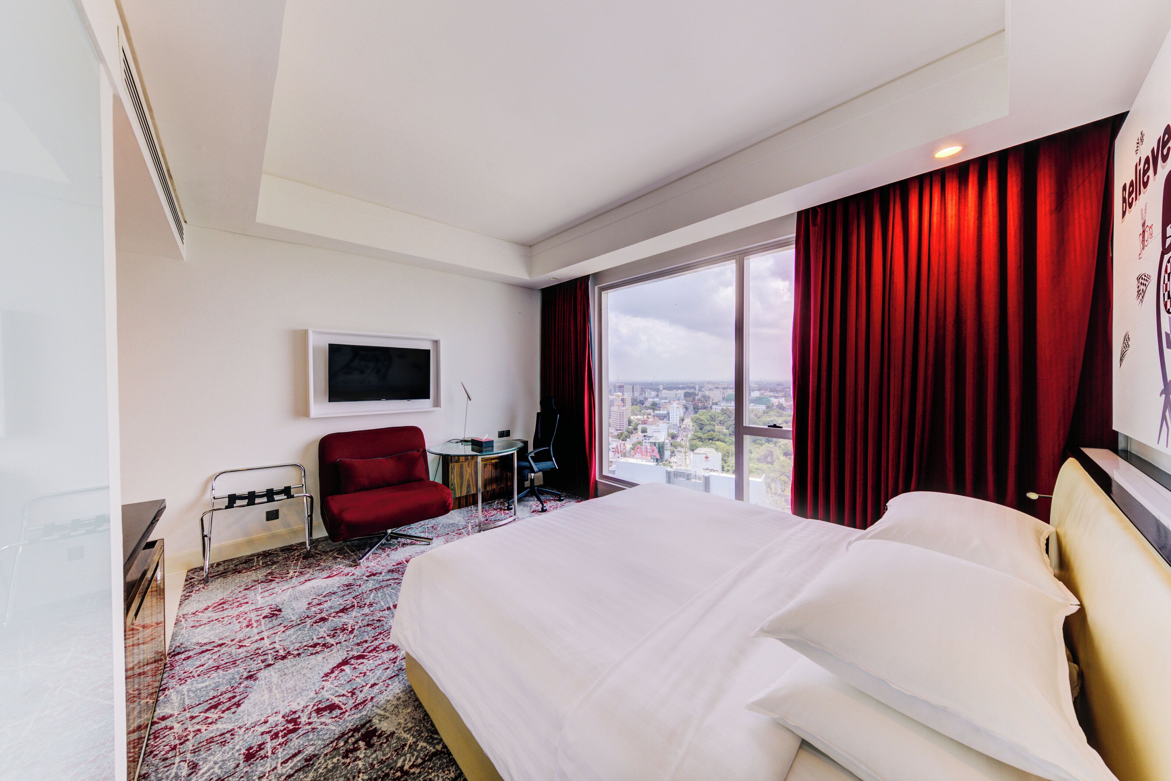 A hotel room with a large bed, a red chair, a TV, and a view of the city.