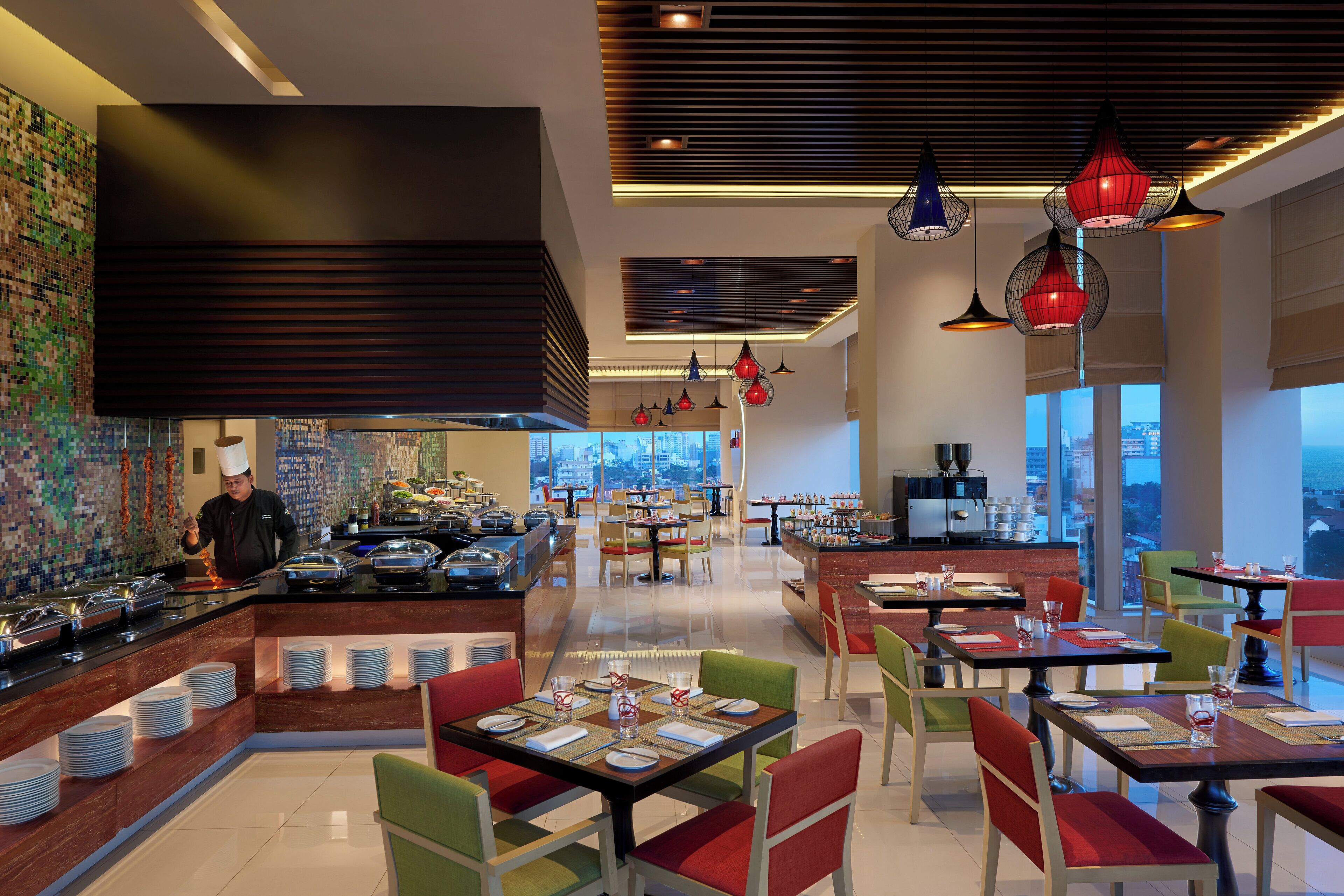 A modern restaurant with a chef preparing food, a buffet area, and dining tables with chairs.