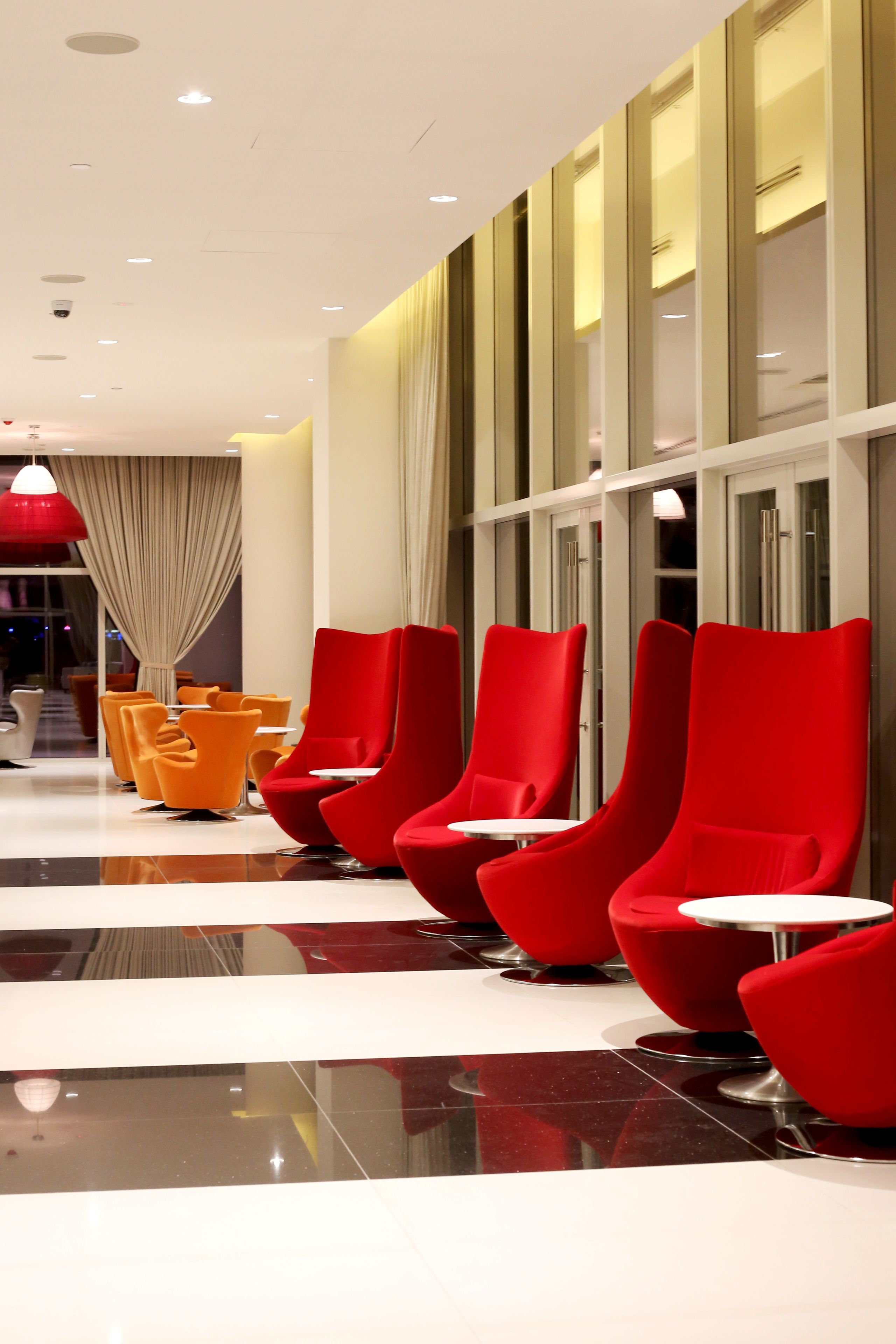 Un hall d’hôtel moderne avec des fauteuils rouges et orange, une cloison en verre et un plafond doté d’un éclairage encastré.