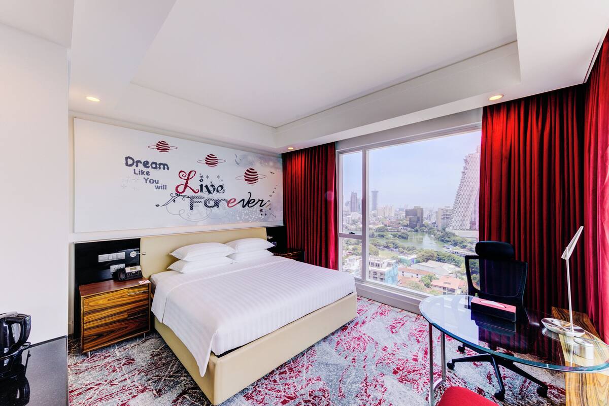 Habitación de hotel moderna con una cama grande, un escritorio con silla, vista a la ciudad y un cuadro motivacional en la pared.