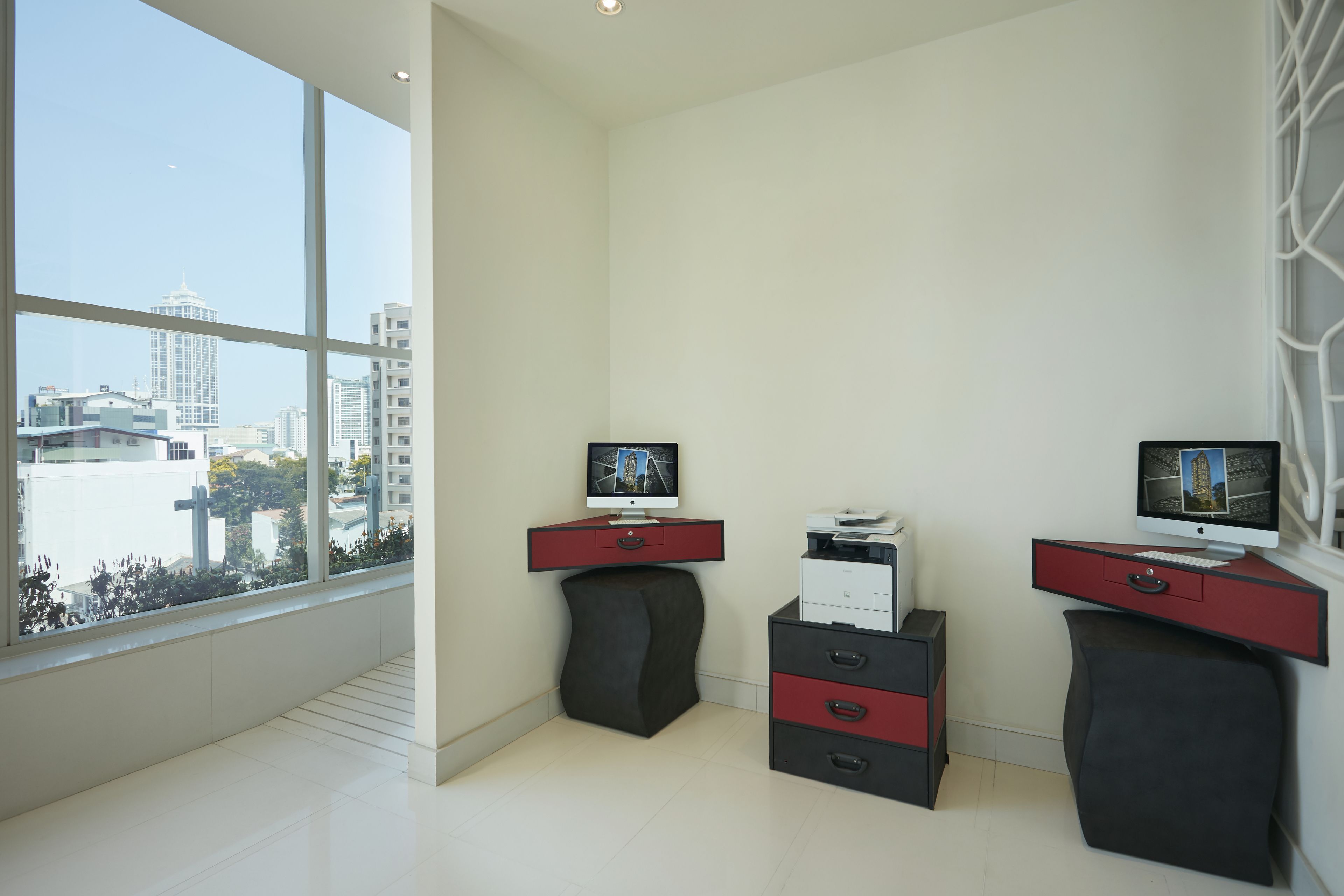 A modern office space with two desks, each with a computer monitor, a printer, and drawers. Large windows offer a view of the cityscape.