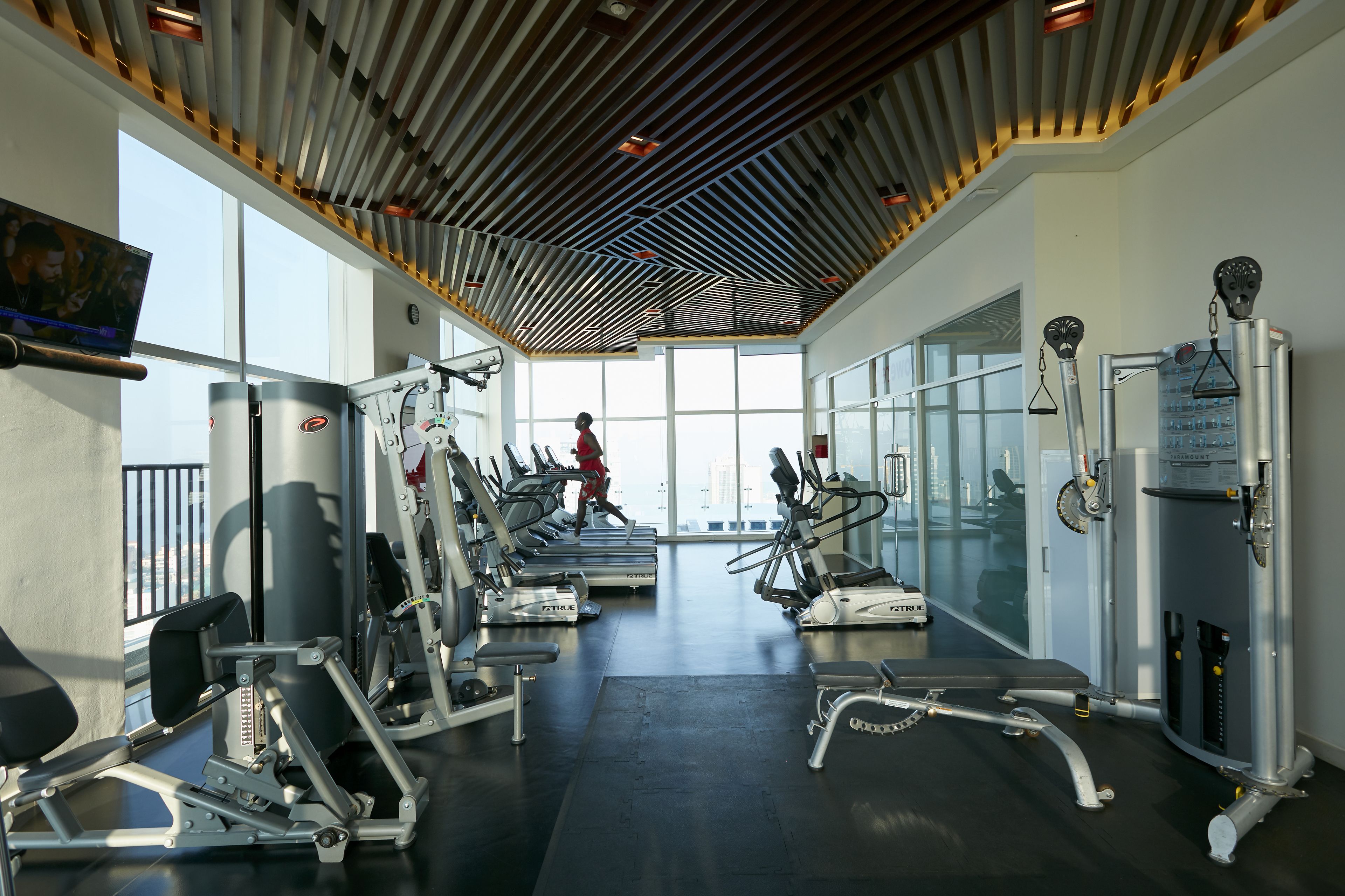 A gym with various exercise machines, large windows, and a high ceiling with a wooden design.