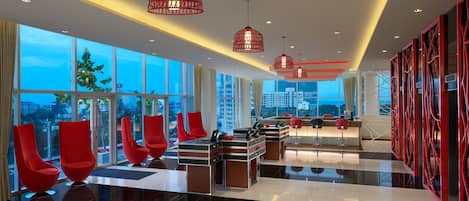 A modern hotel lobby with large windows, red chairs, a reception desk, and red pendant lights.