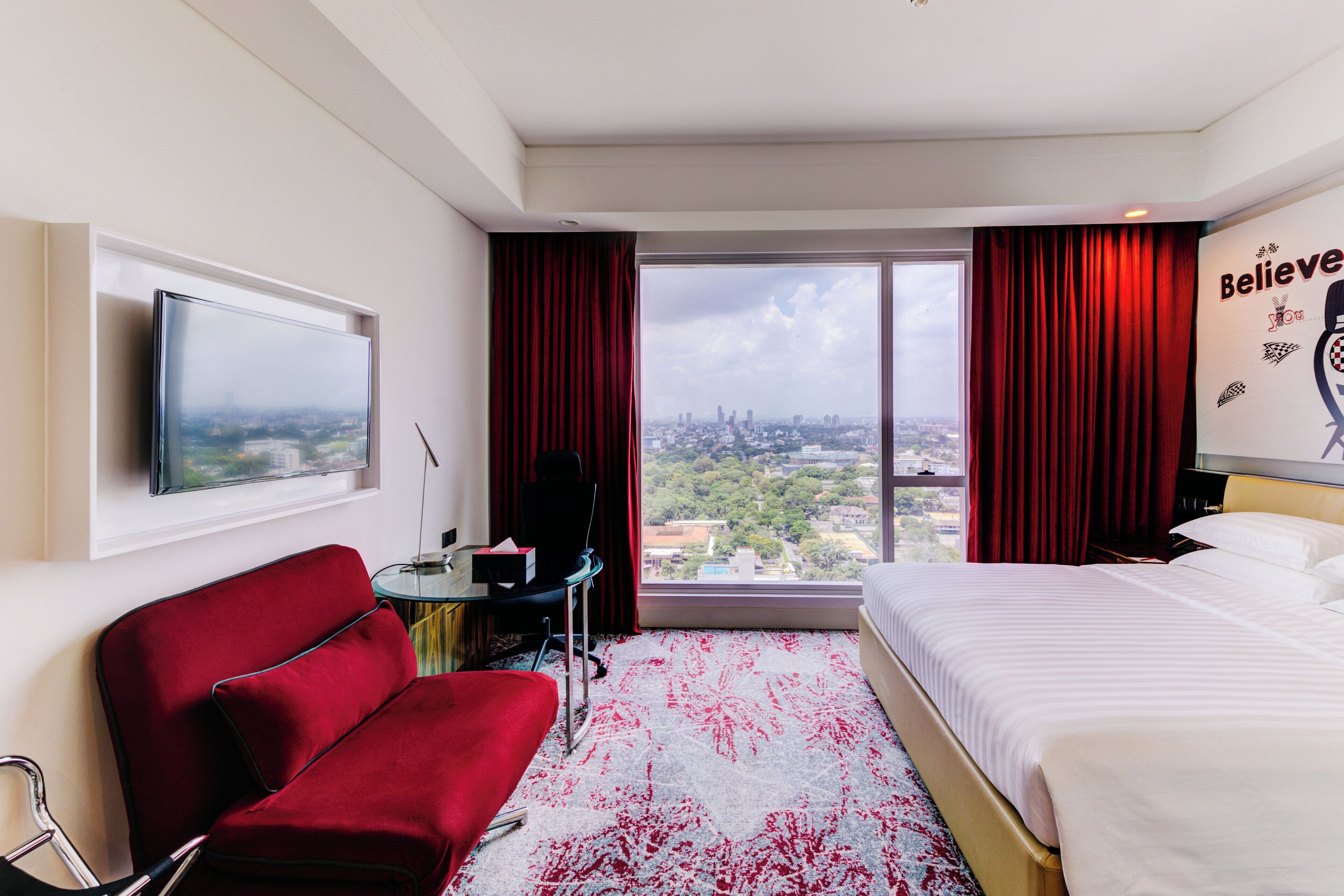Habitación de hotel con cama, un sillón rojo, un escritorio con silla, televisor en la pared y un amplio ventanal con cortinas rojas.