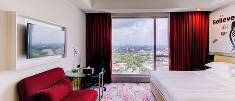 Habitación de hotel con cama, un sillón rojo, un escritorio con silla, televisor en la pared y un amplio ventanal con cortinas rojas.
