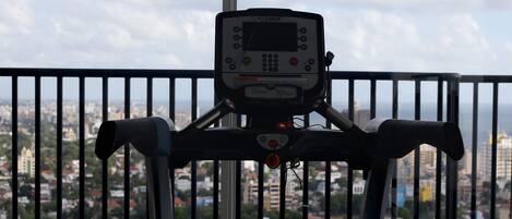 Una bicicleta estática con pantalla digital, ubicada frente a una ventana con vista a la ciudad.