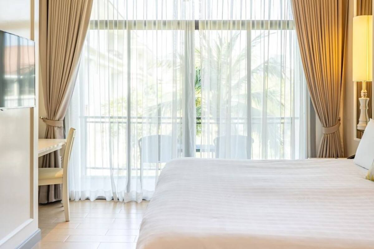 Une chambre d’hôtel avec un grand lit, un téléviseur fixé au mur, une chaise et une vue sur les palmiers à travers de fines rideaux.