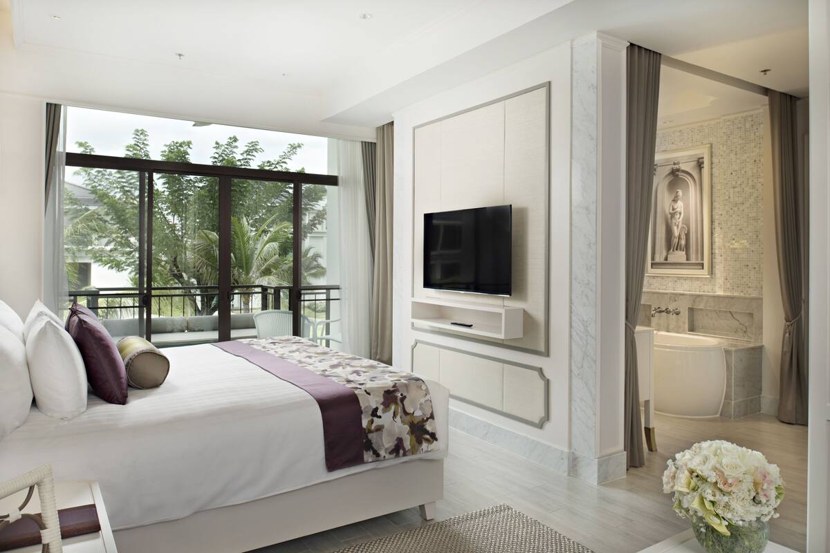 Une chambre d’hôtel moderne dotée d’un grand lit, d’une télévision, d’une salle de bain avec baignoire et d’un balcon offrant une vue sur les arbres.