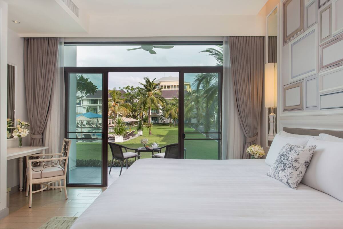 Une chambre d’hôtel avec un grand lit, un bureau avec une chaise, une petite table avec des chaises, et une vue sur un jardin et des bâtiments à l’extérieur.