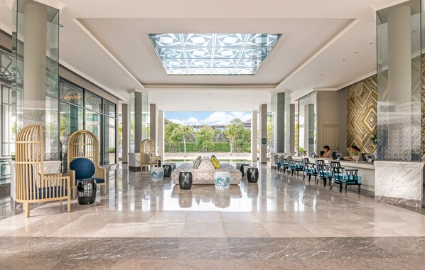 Un lobby espacioso con techo alto, ventanas grandes y una zona de estar con sillas y sofá.