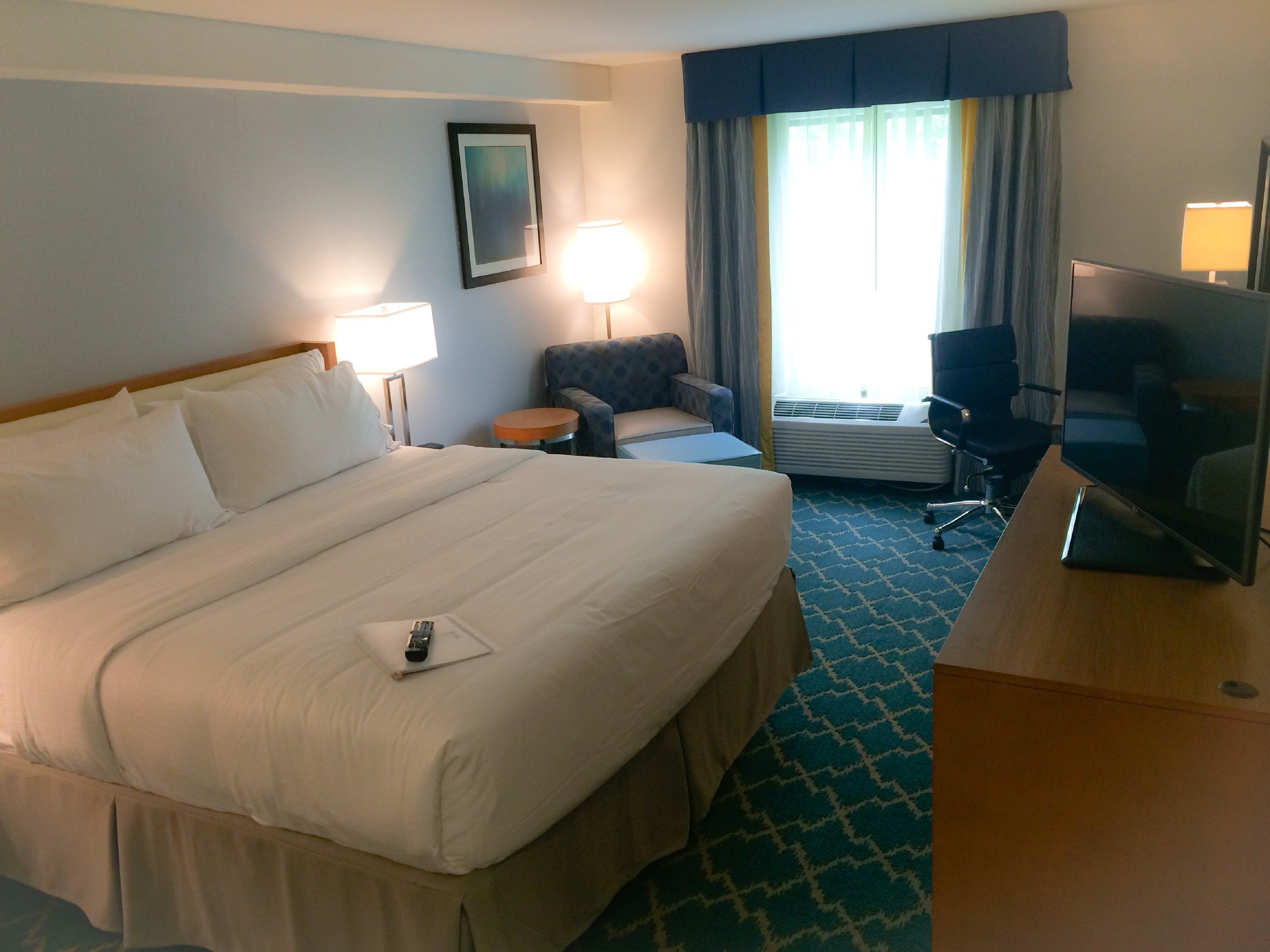 A hotel room with a large bed, a desk with a television, a chair, a lamp, and a window with curtains.