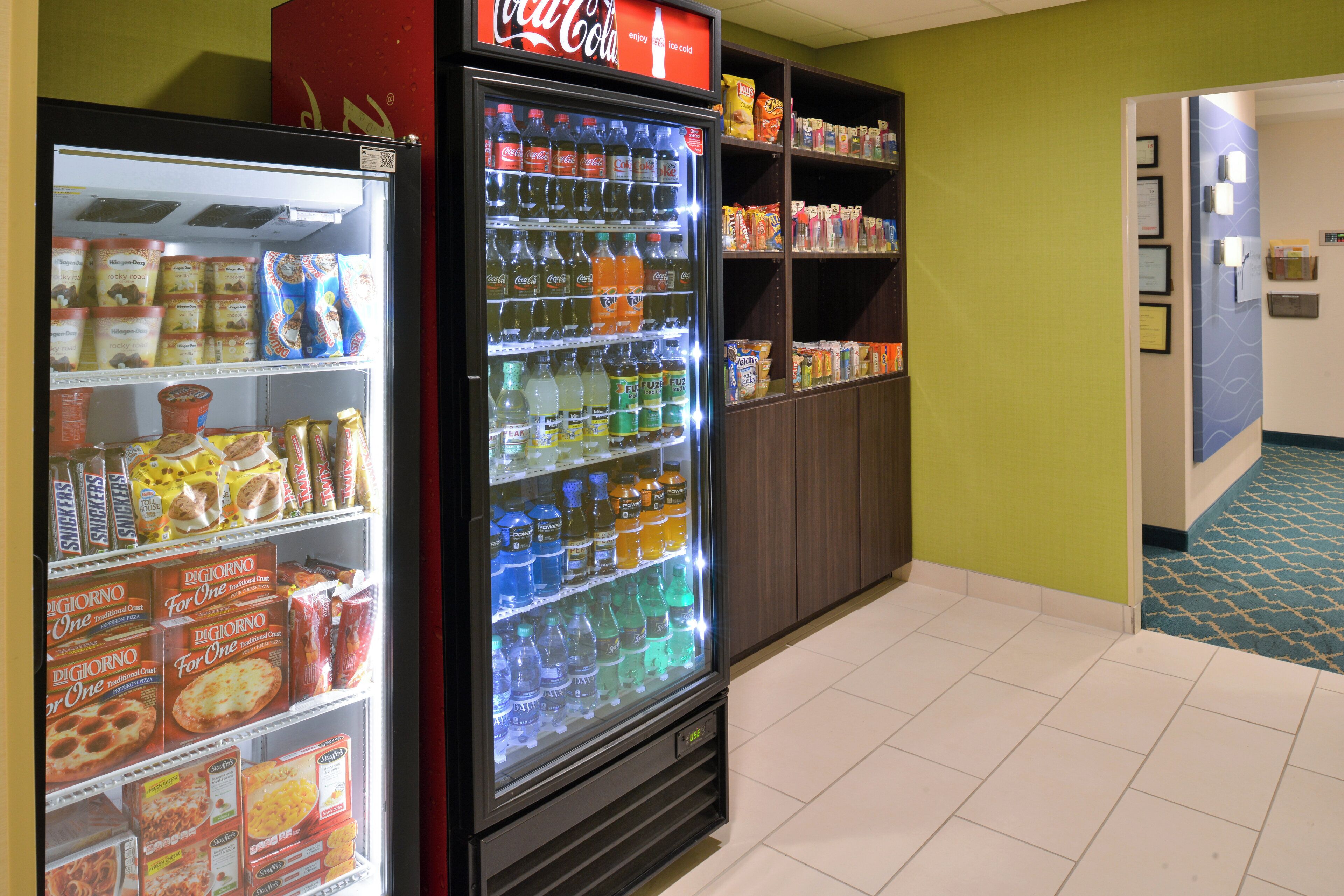 Ein Getränkeautomat mit verschiedenen Snacks und Getränken, ein Kühlschrank mit Eis und Tiefkühlkost sowie Regale voller Snacks und Getränke.