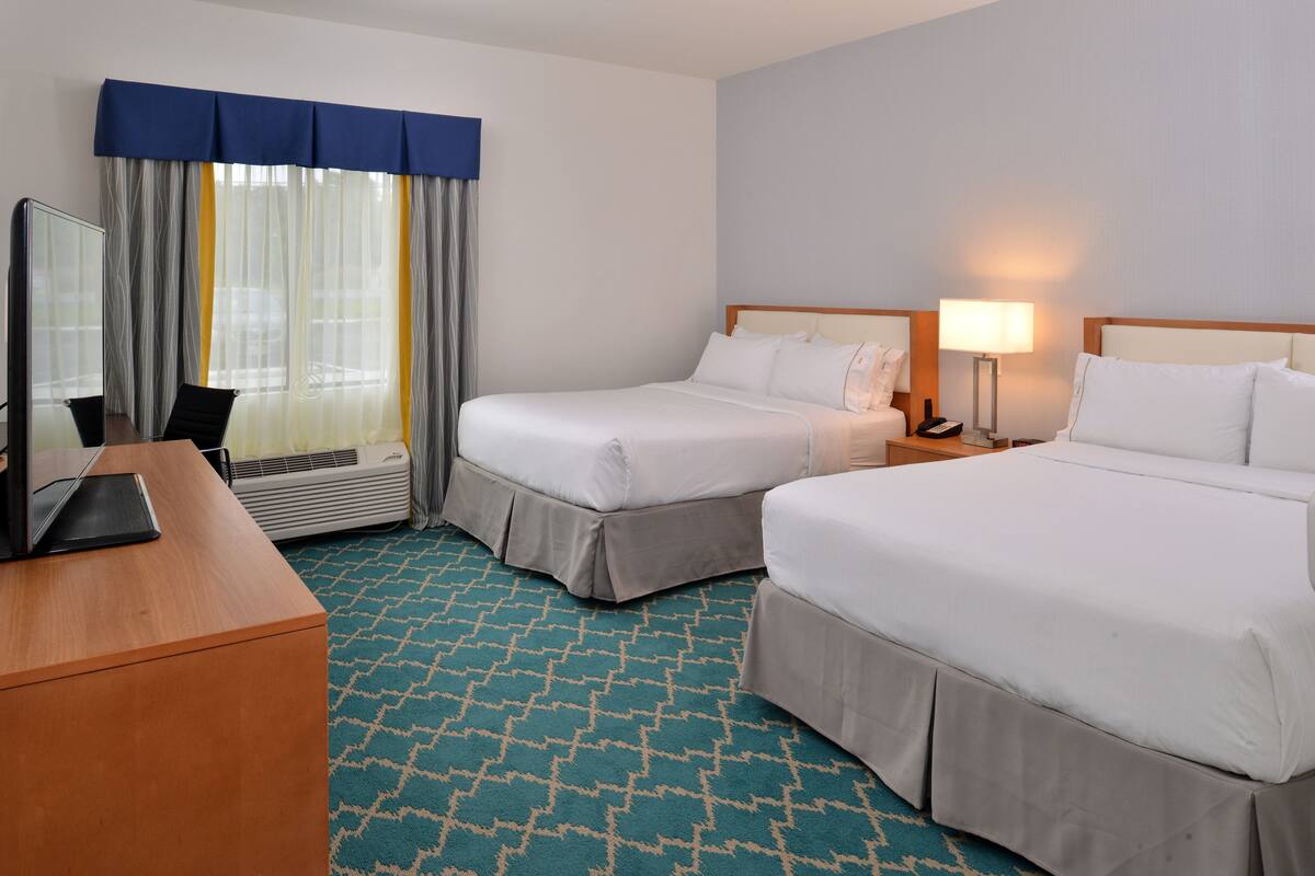 A hotel room with two beds, a desk with a laptop, a desk lamp, and a window with curtains.