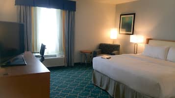Habitación de hotel con cama, escritorio con televisor, silla, lámpara y ventana con cortinas.