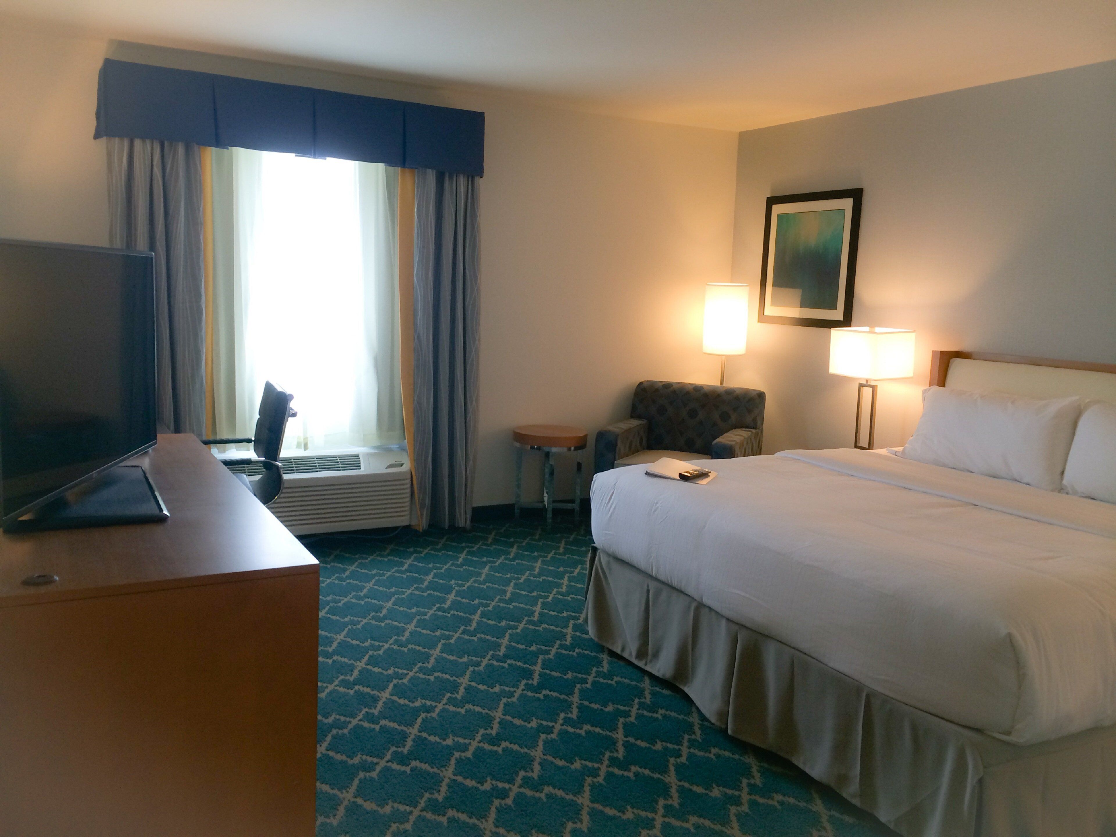 Habitación de hotel con una cama, un escritorio con televisor, una silla, una lámpara y un cuadro en la pared.