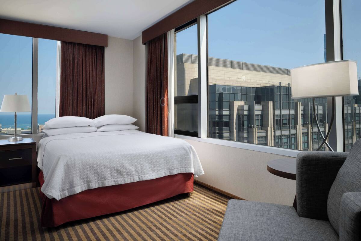 Une chambre d’hôtel avec un grand lit, un bureau avec une lampe, une chaise et une vue sur un immeuble moderne à travers de grandes fenêtres.