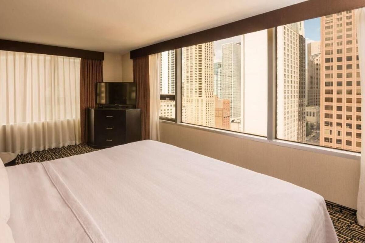 Une chambre d’hôtel avec un lit, une télévision, une commode et une grande fenêtre donnant sur la ville.
