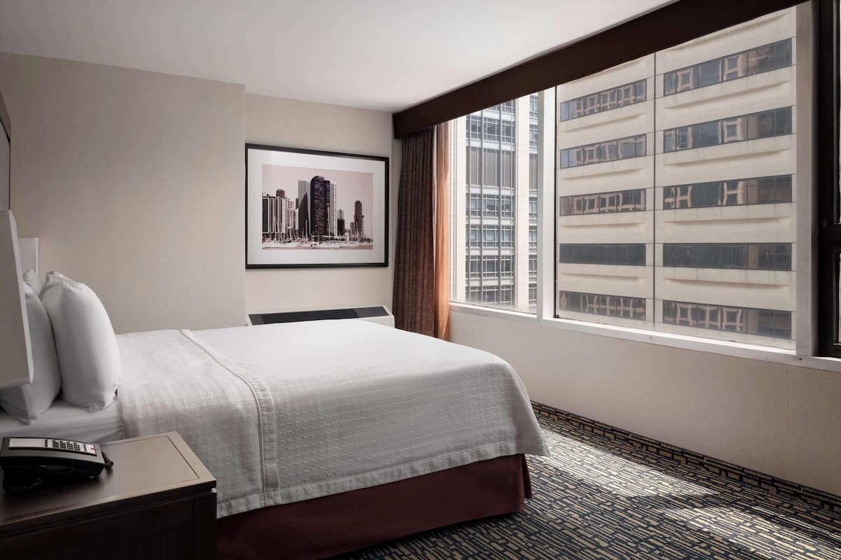 Une chambre d’hôtel avec un grand lit, un tableau représentant une vue de la ville encadré au mur, une fenêtre donnant sur des immeubles de grande hauteur et un téléphone sur la table de chevet.