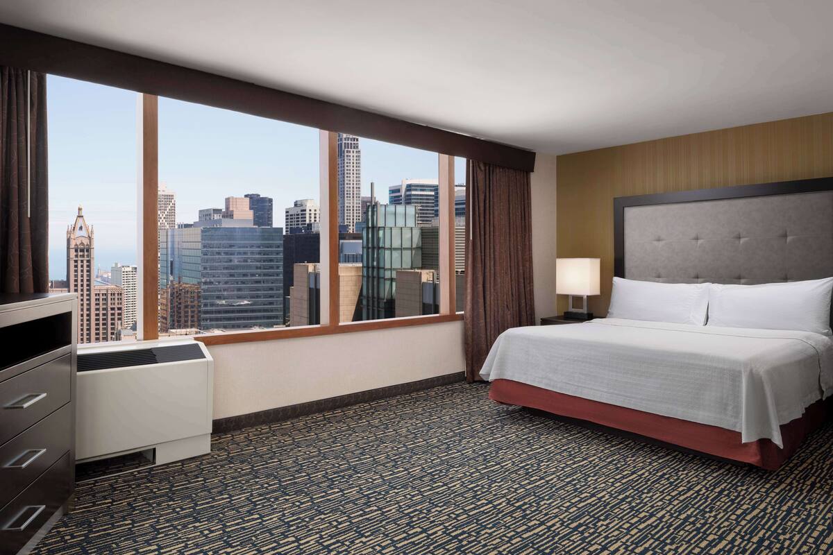 Une chambre d’hôtel dotée d’une grande fenêtre donnant sur la ville, d’un lit avec une tête de lit grise, d’une table de chevet avec une lampe et d’une commode.