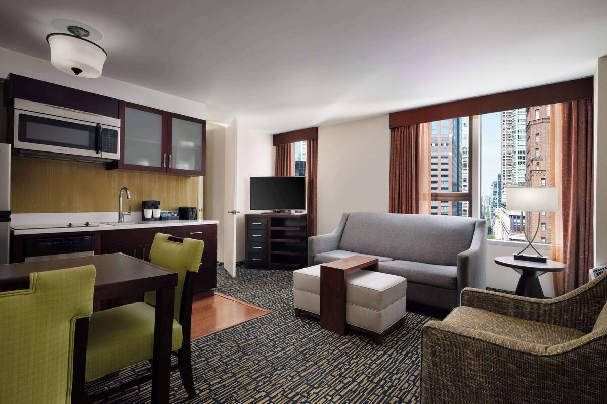 Une chambre d’hôtel avec une kitchenette, un canapé gris, une table à manger avec des chaises et une vue sur la ville grâce à de grandes fenêtres.