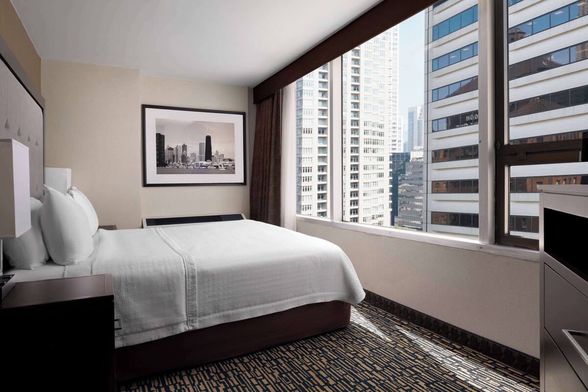 Une chambre d’hôtel avec un grand lit, un tableau représentant une vue sur la ville encadré au mur et une vue sur de hauts immeubles par la fenêtre.