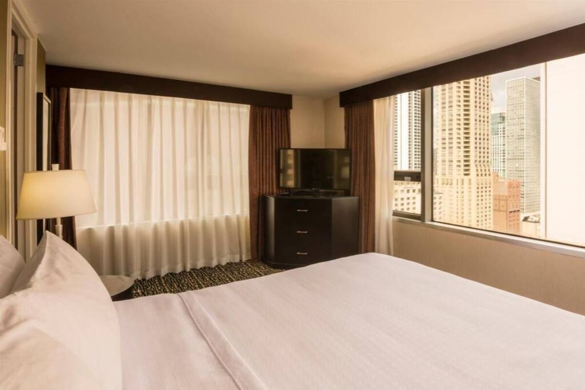 Une chambre d’hôtel équipée d’un lit, d’une télévision, d’une commode et d’une fenêtre donnant sur la ville.