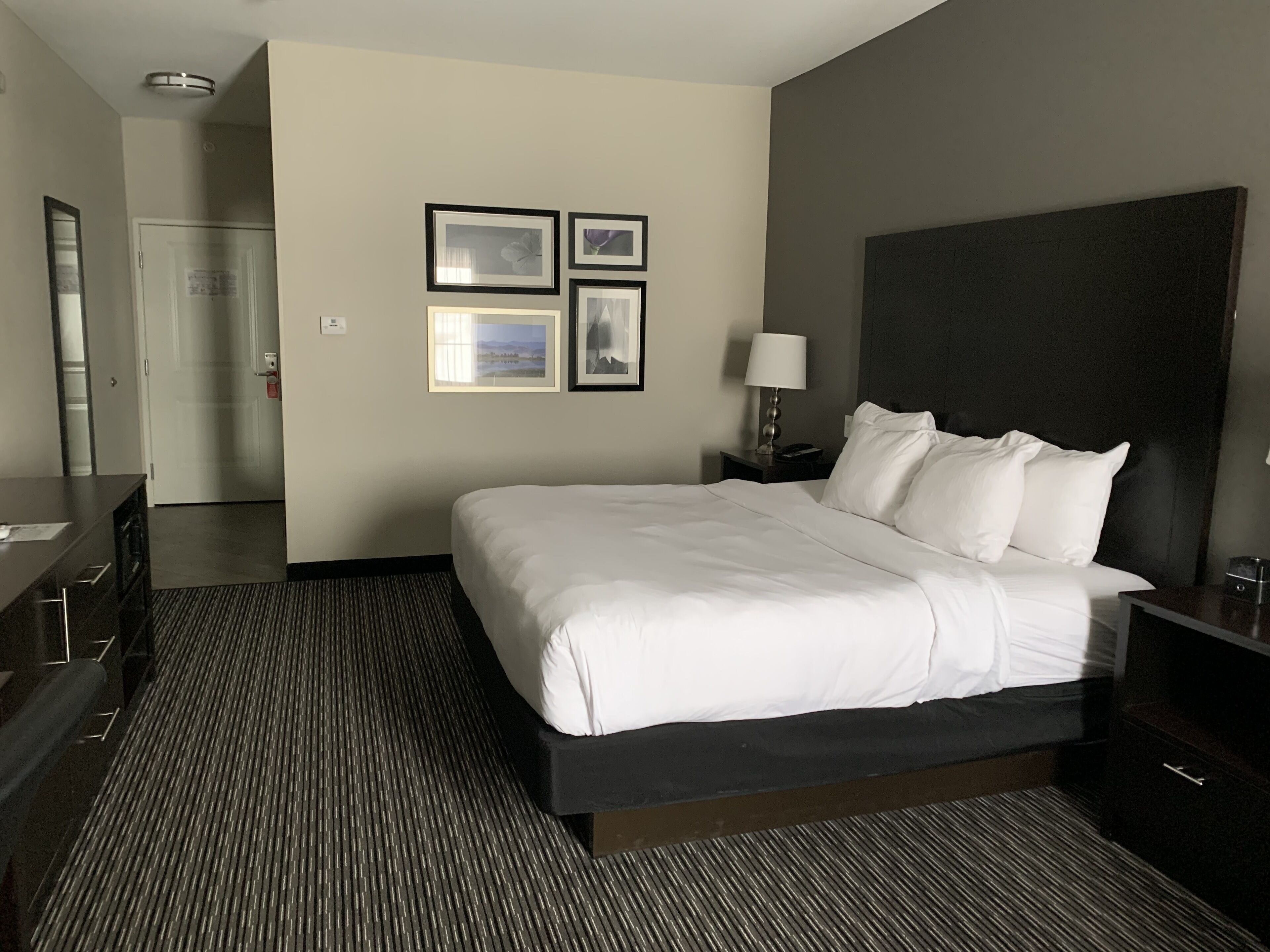 A hotel room with a large bed, a bedside table with a lamp, a dresser, and a mirror. There are framed pictures on the wall and a door leading to another room.