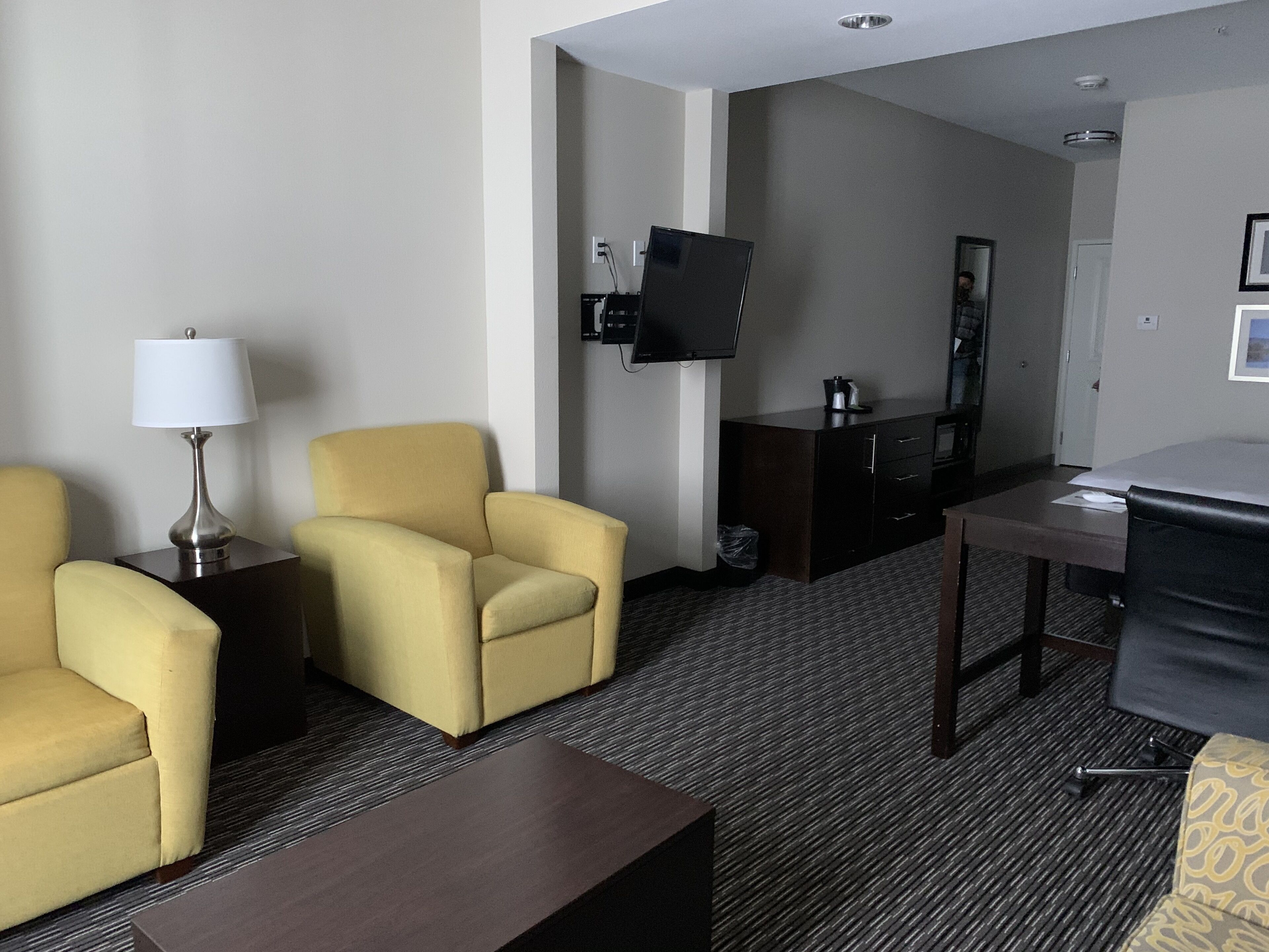 A hotel room with a sofa, armchair, a small table, a TV mounted on the wall, and a desk.