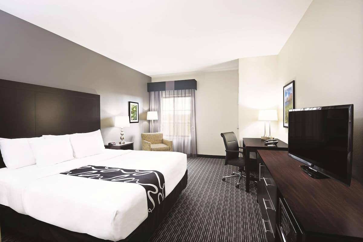 A hotel room with a large bed, a desk with a flat-screen TV, a chair, a lamp, and a window with curtains.