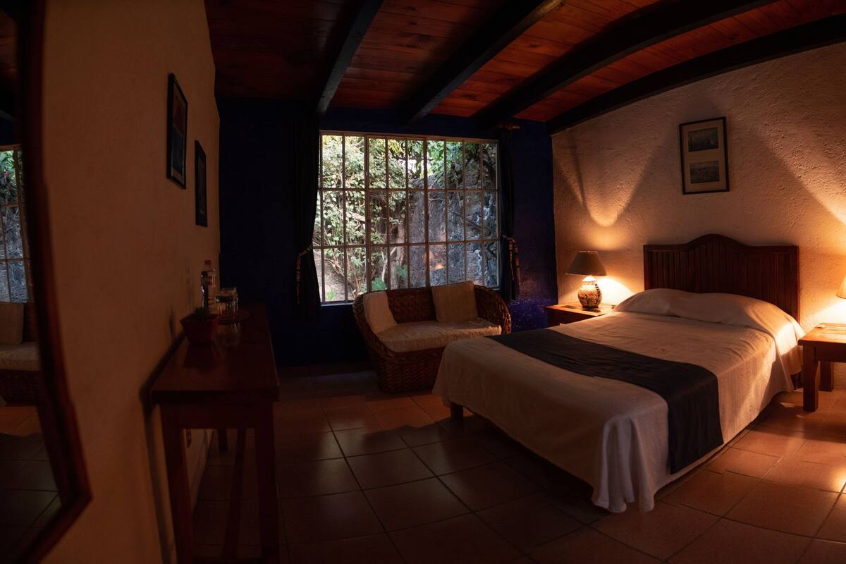 A cozy bedroom with a large bed, two bedside tables with lamps, a wicker chair, and a window with a view of trees.