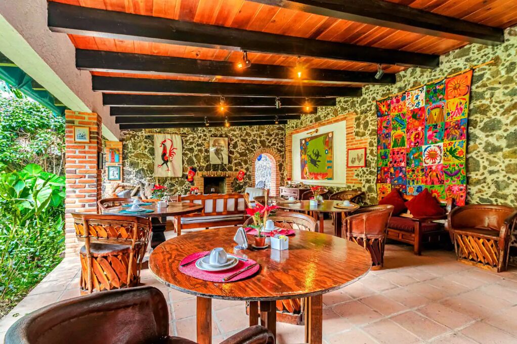 An outdoor dining area with rustic wooden tables and chairs, stone walls, and a colorful mural.
