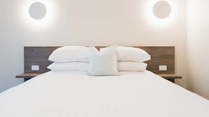 A neatly made bed with white linens and pillows, two bedside tables with light switches, and two wall-mounted circular lights. - West End Motor Lodge (Orange)