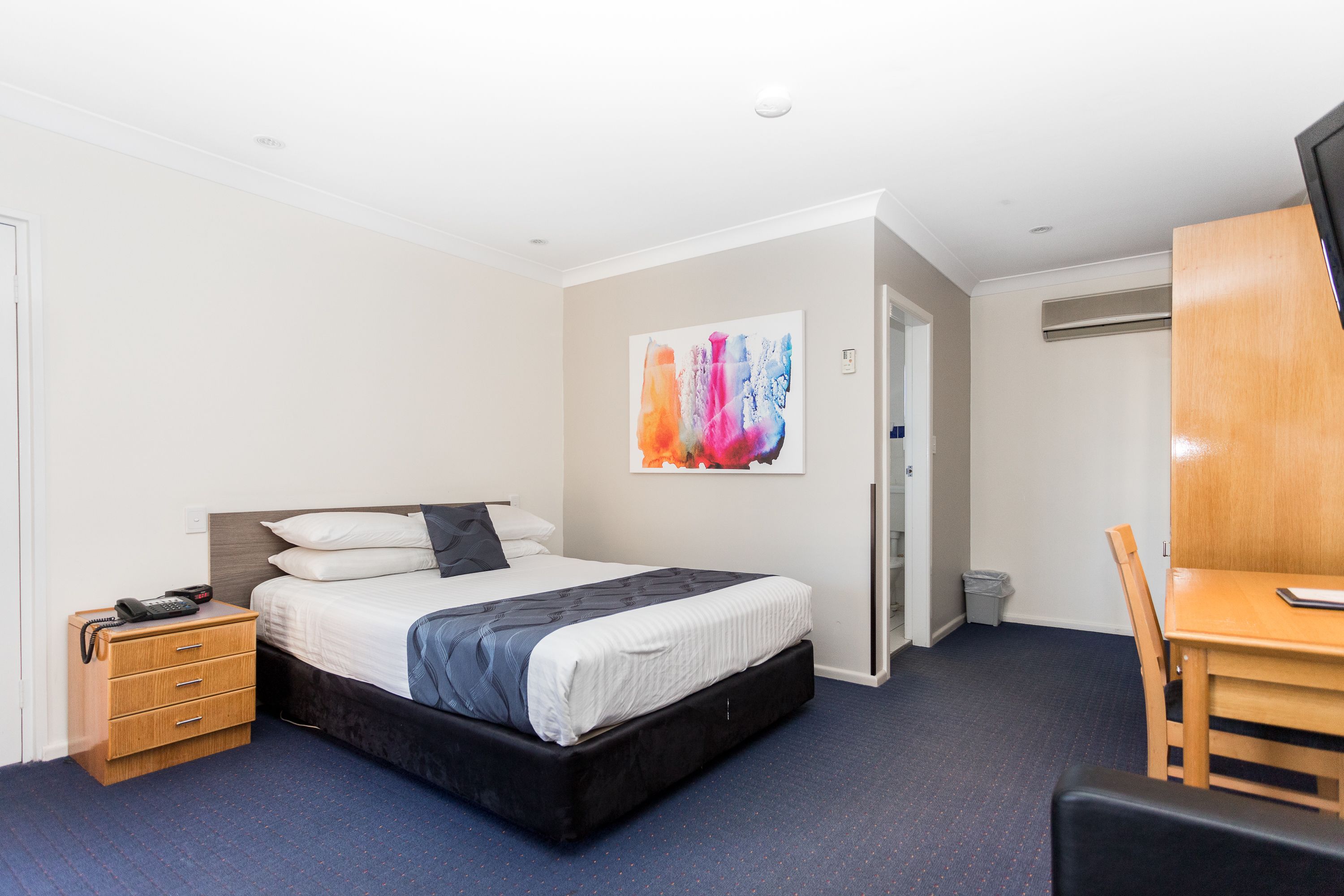 A hotel room with a bed, bedside table, chair, desk, and a painting on the wall.