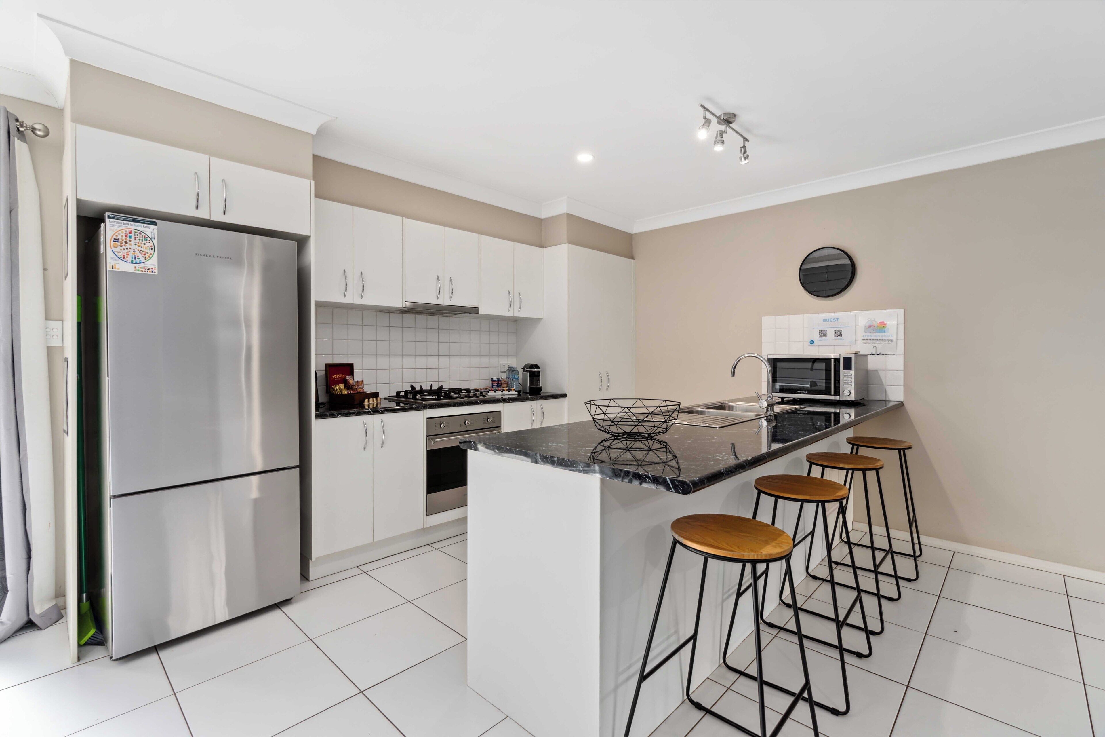 A modern kitchen with a stainless steel refrigerator, a gas stove, and a microwave. There are four bar stools and a large island with a marble countertop.