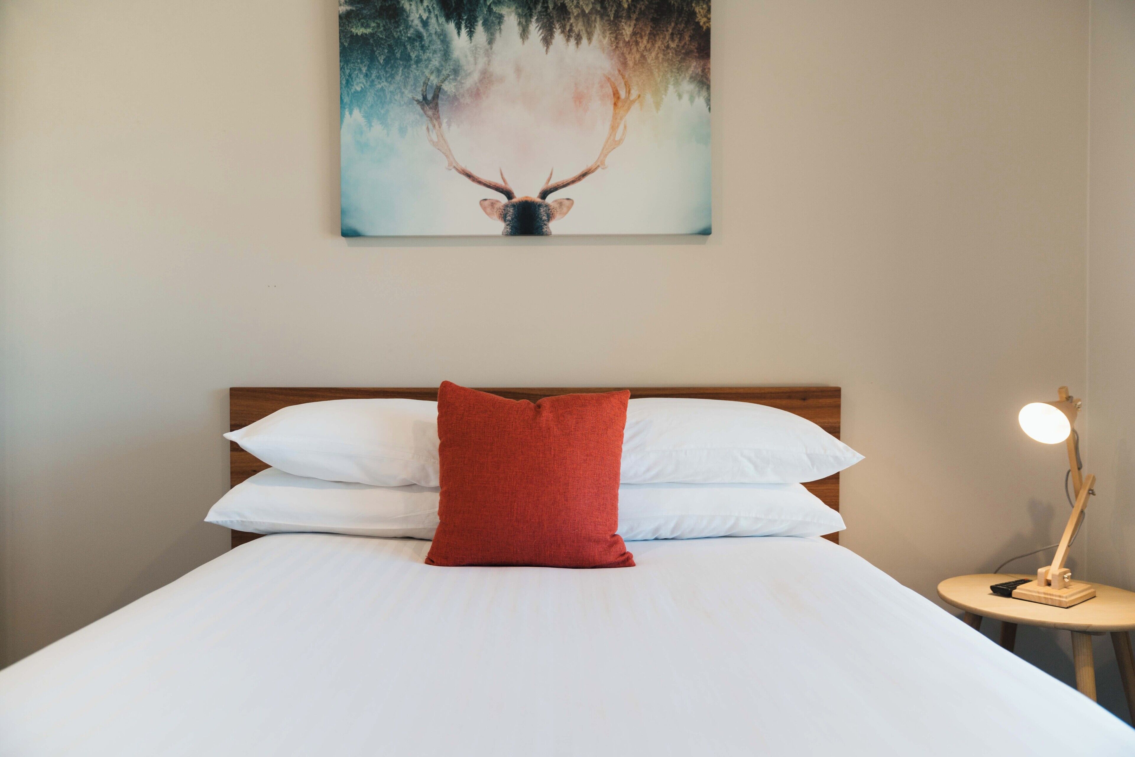 A neatly made bed with white and red pillows, a wooden headboard, and a painting of a deer above it.